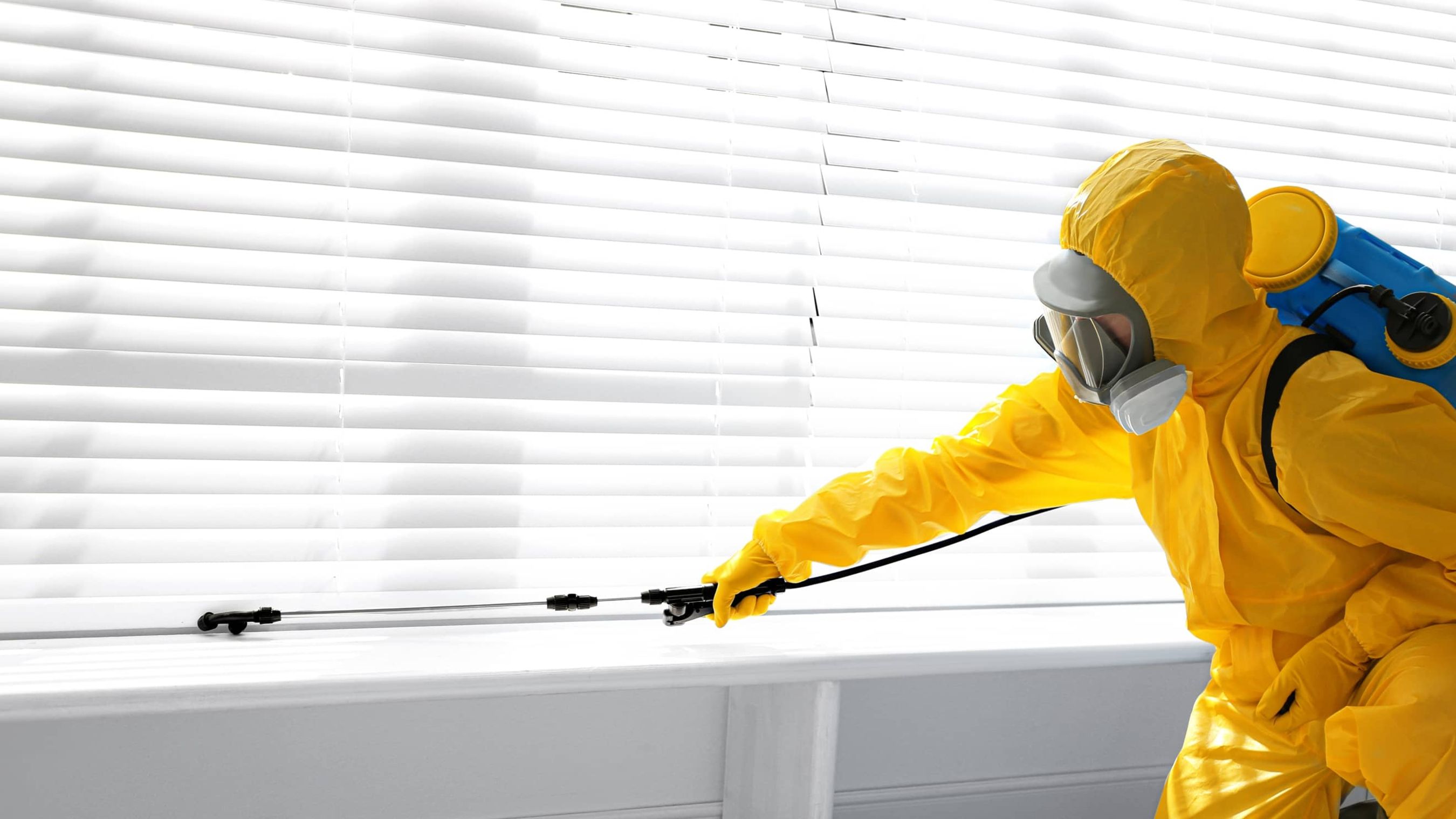 Person in gelbem Schutzanzug und Atemmaske sprüht mit einer langen Düse vor einem Fenster mit weißen Jalousien